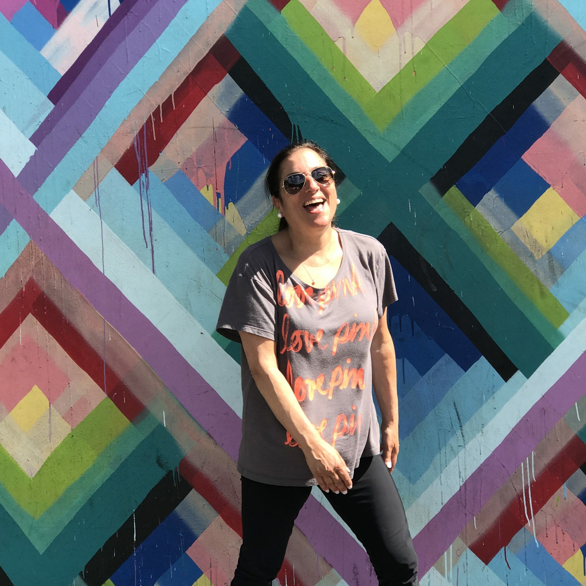 A photo of Shelley who has brown hair pulled back, and wearing sunglasses in front of a brightly colored background.